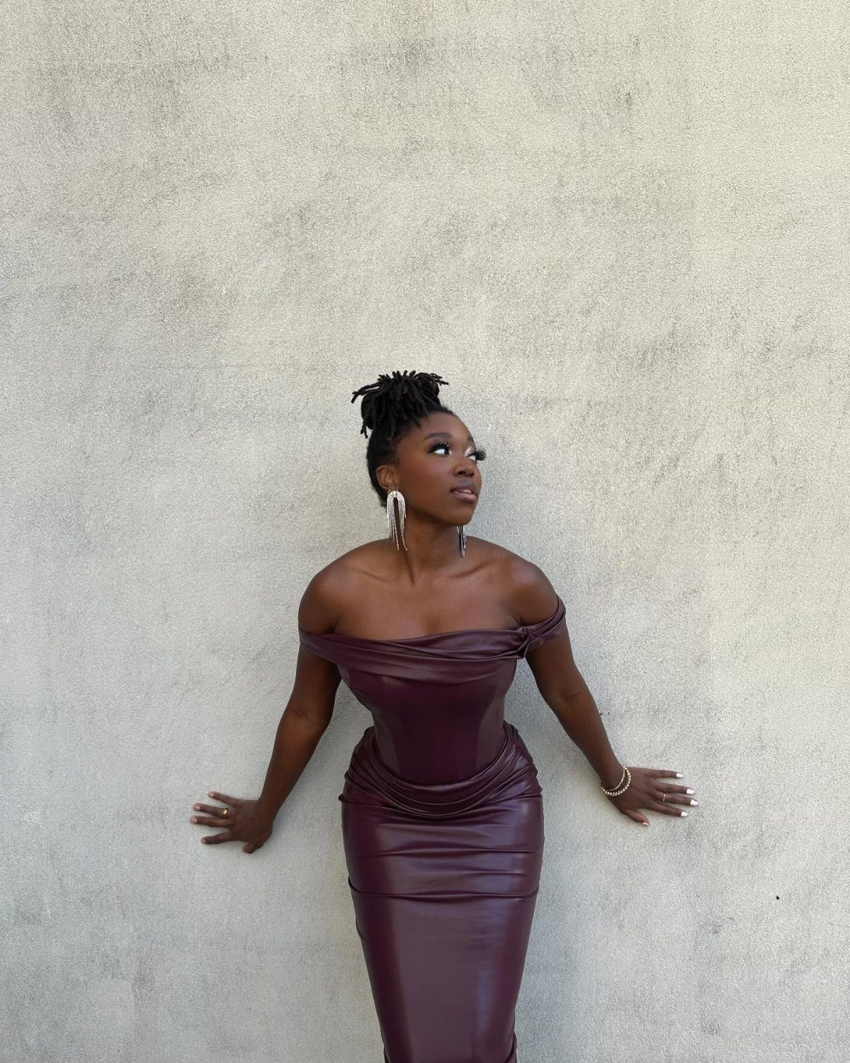 Woman in a burgundy off-the-shoulder dress posing against a textured grey wall.
