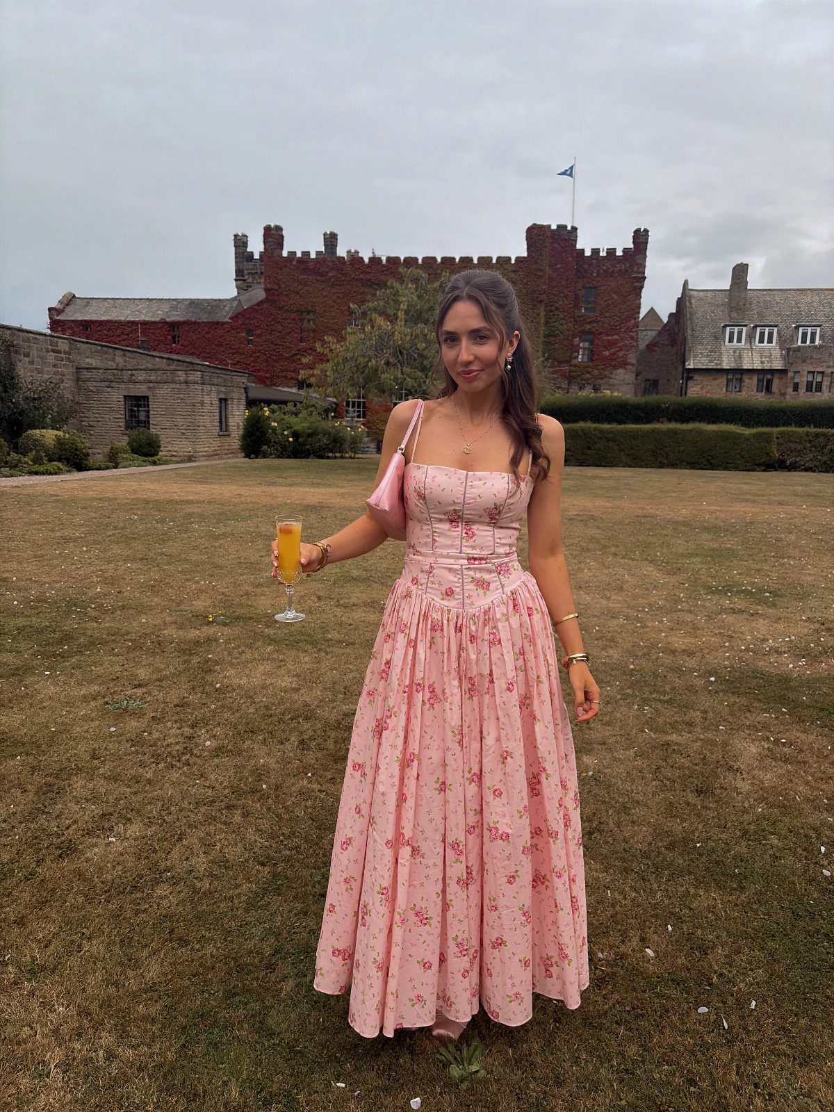 Woman in a pink floral corset maxi dress standing on a lawn before a castle.