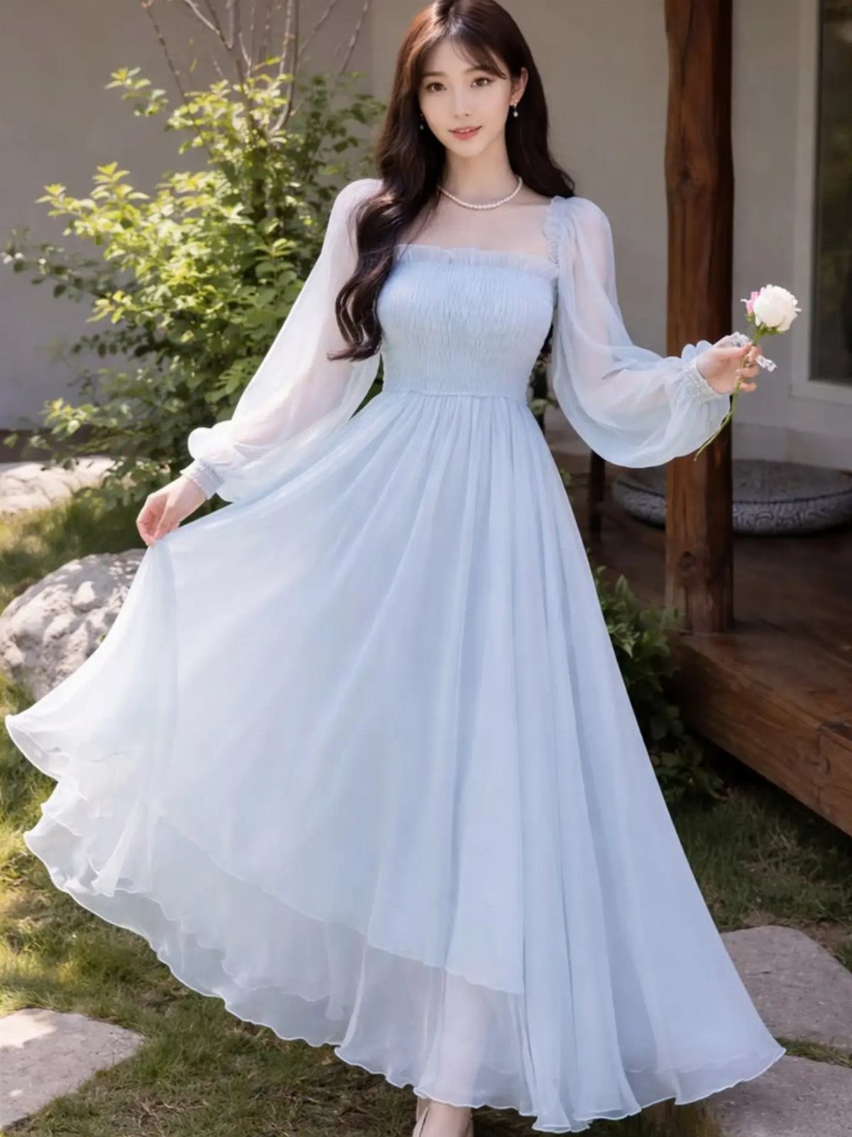 A woman in a flowing light blue long-sleeved maxi dress standing in a garden holding a rose.
