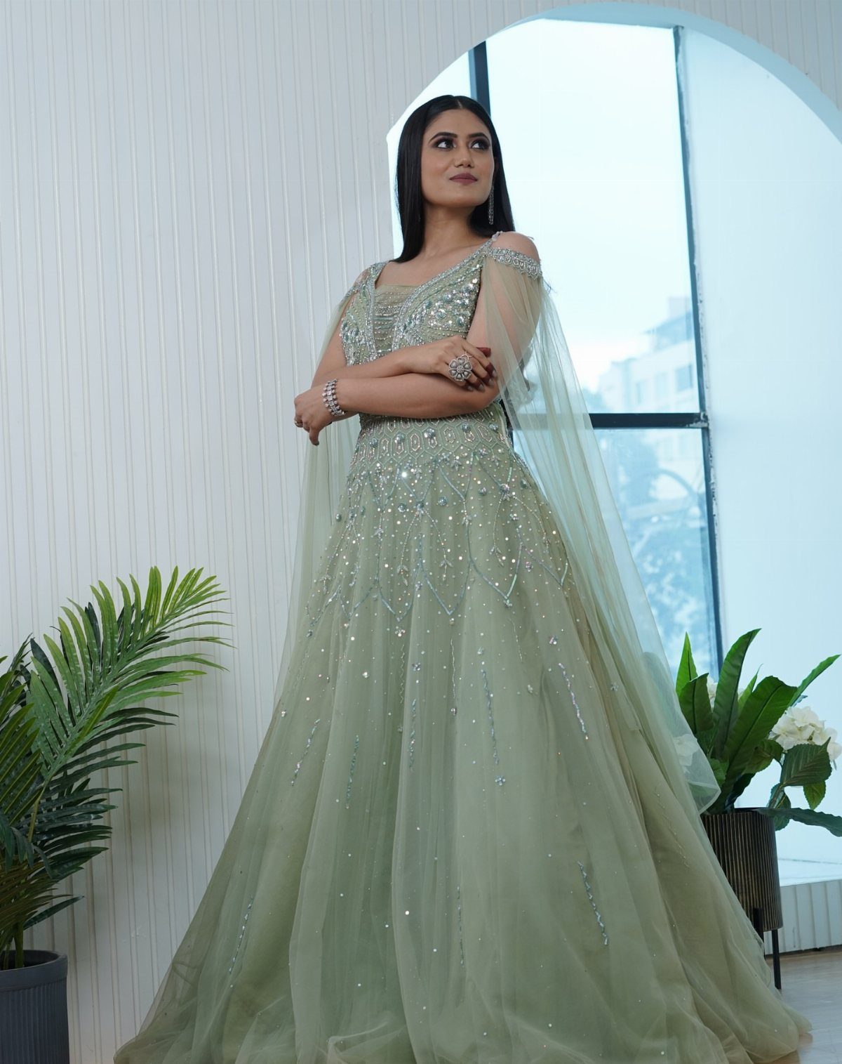 Woman in a light green sequined floor-length gown standing near an arched window.