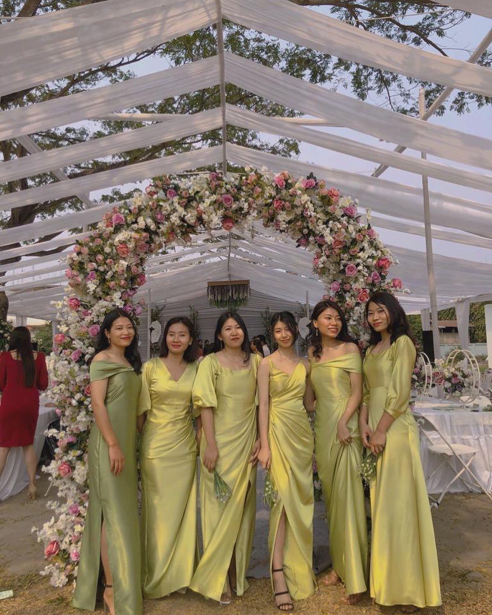Six women posing in light green satin formal dresses under a floral archway.