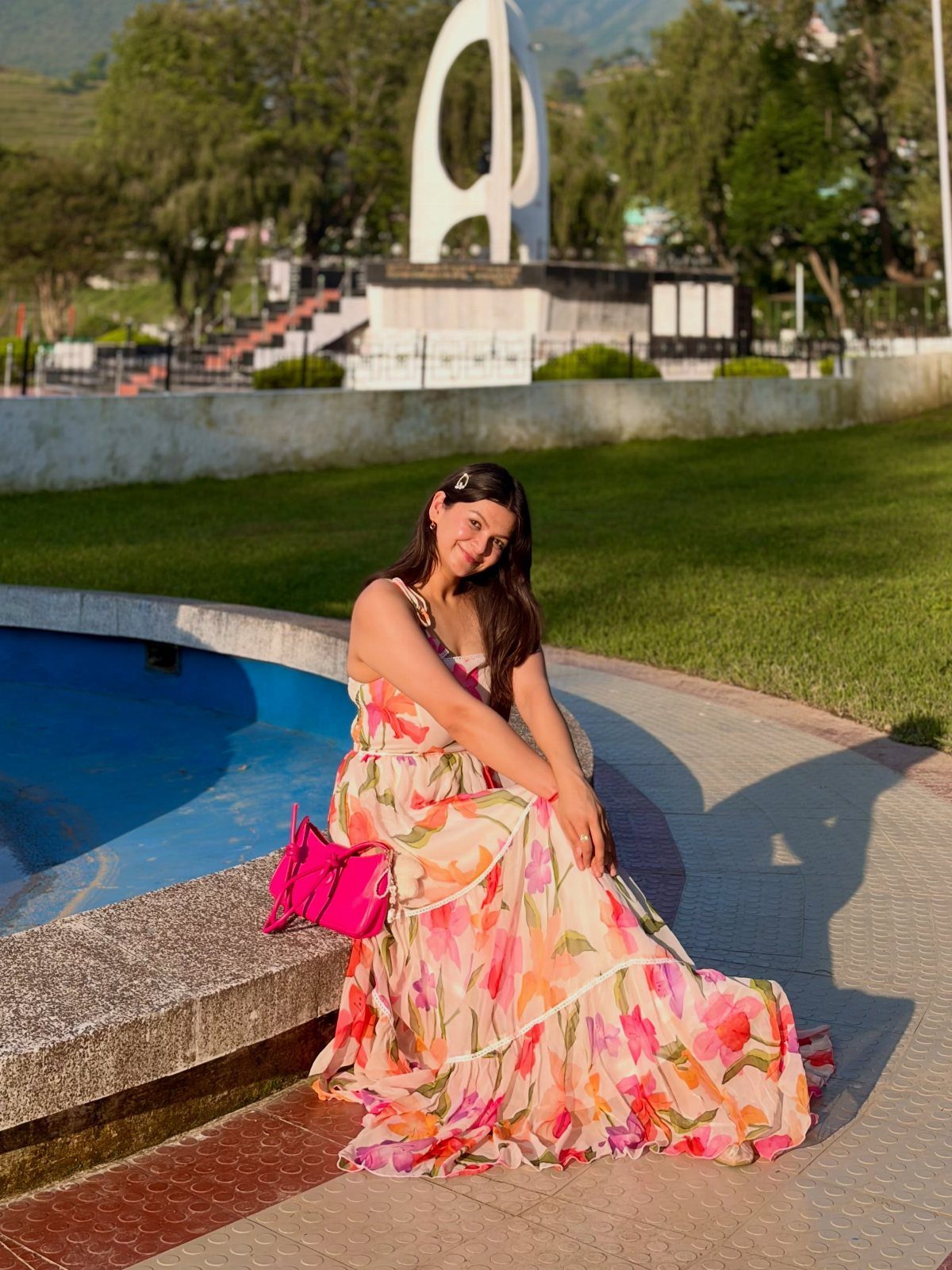 Woman in a pink floral print maxi dress sitting by a blue pool outdoors.