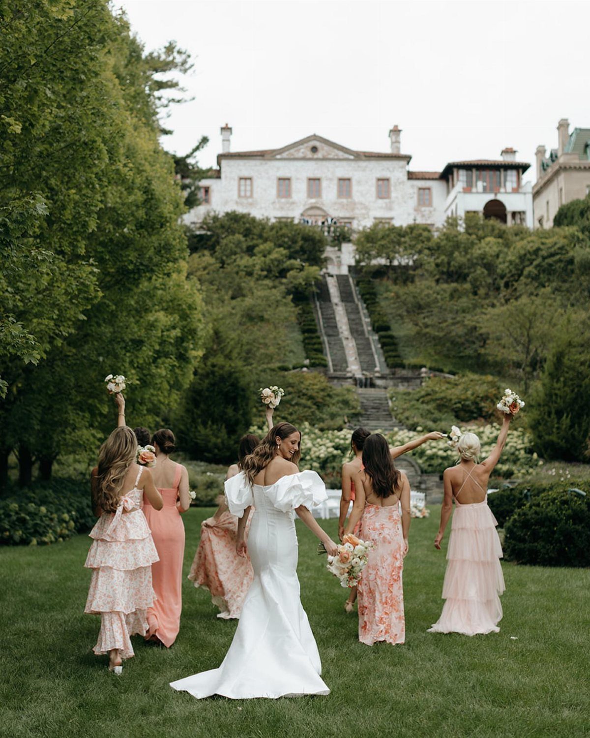 Bridesmaids in pink and peach dresses walk with a bride in a white gown through a garden.