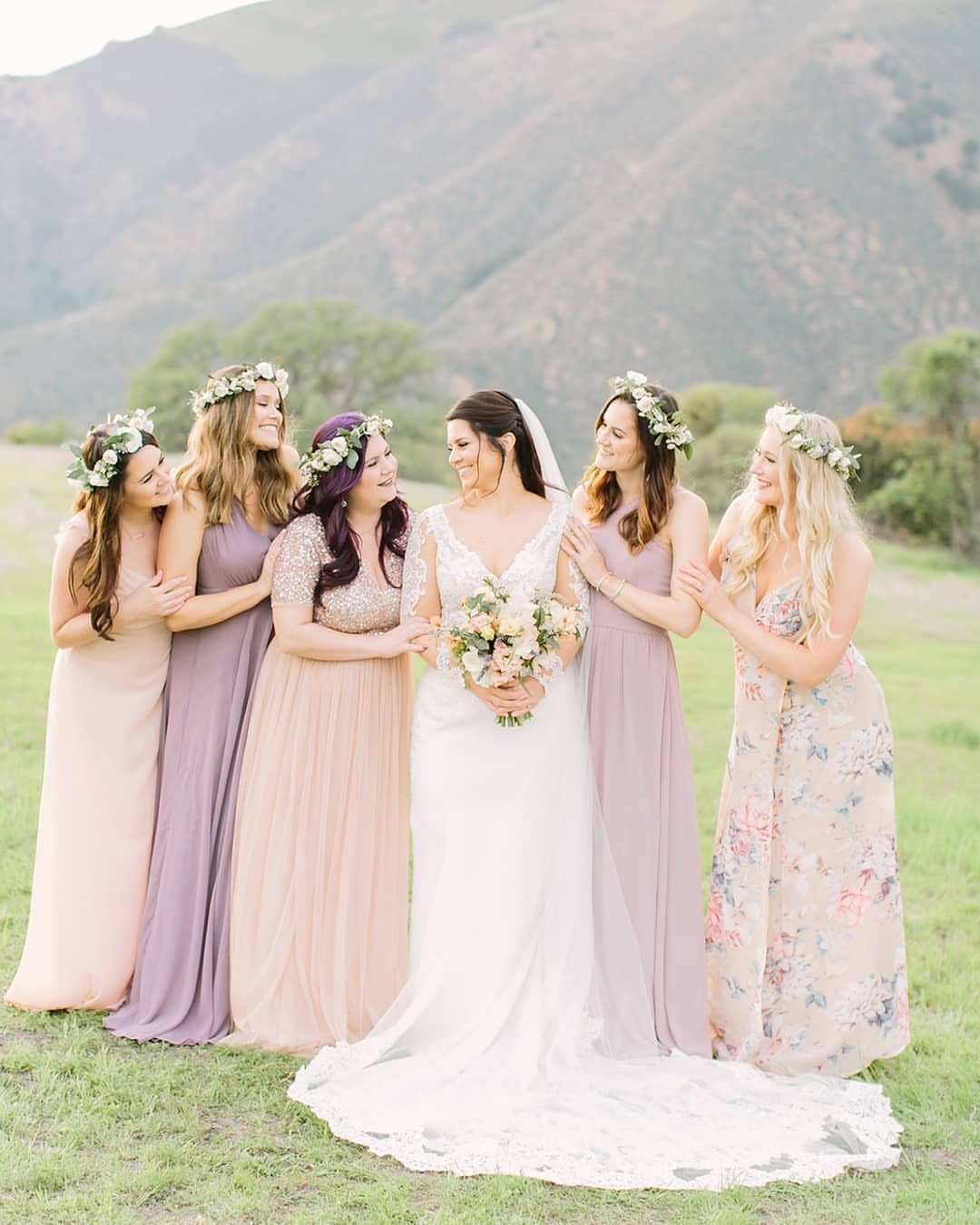 Bride in white lace surrounded by bridesmaids wearing various colored and patterned long dresses outdoors.