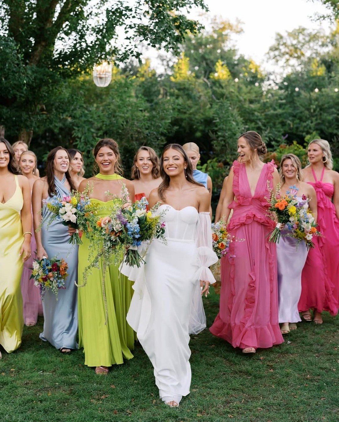 Bridesmaids in colorful dresses walking through a lush green garden with the bride in white.