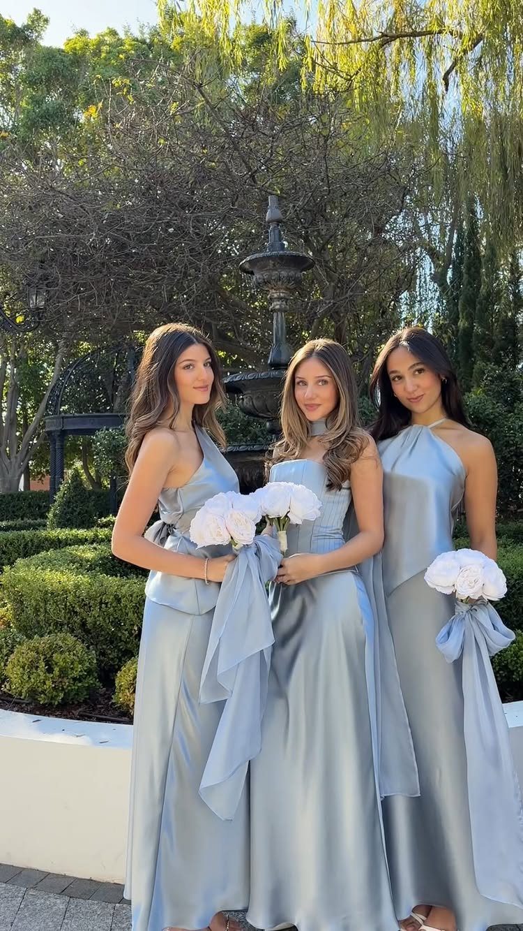 Three bridesmaids in different styles of dusty blue satin dresses holding white bouquets outdoors.