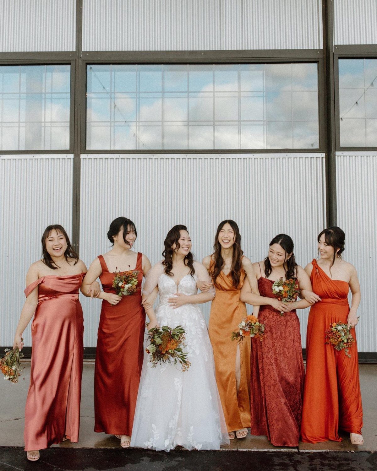 Bridesmaids in various shades of orange and terracotta dresses standing with a bride in white.