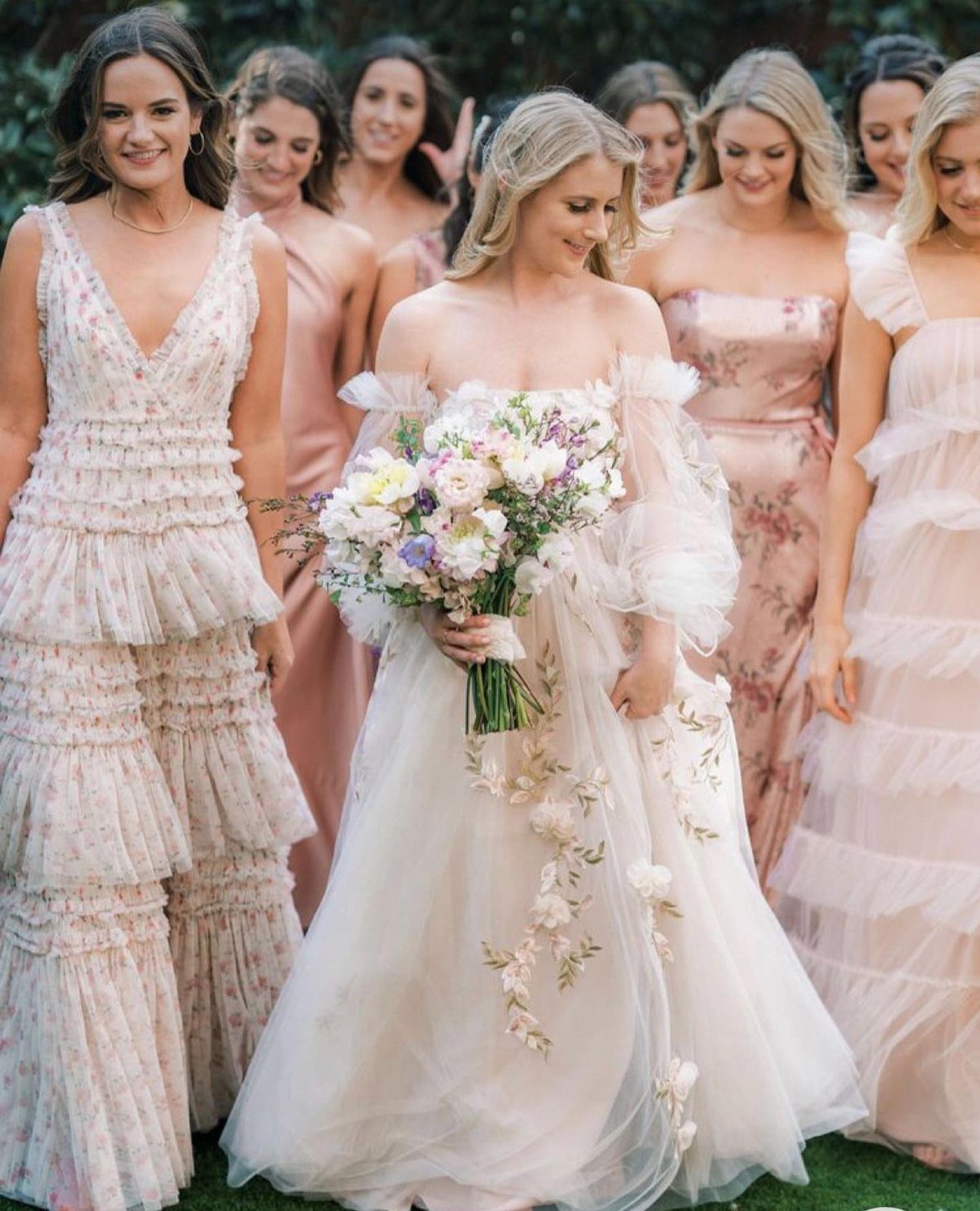 A bride in a white floral gown walks with bridesmaids in various pink and cream dresses.