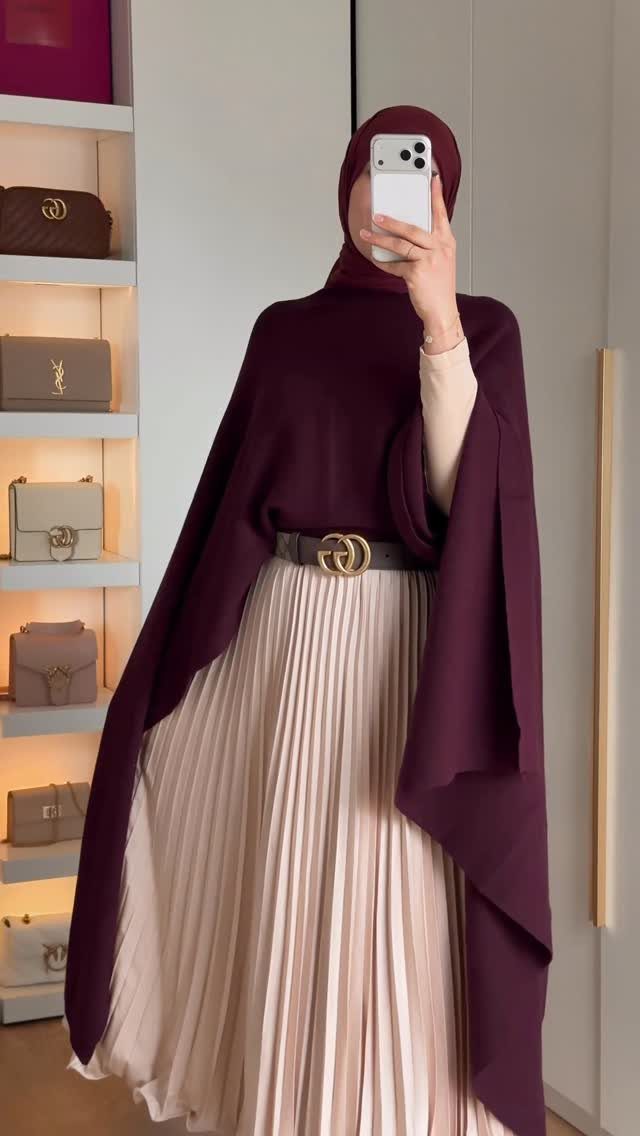 A person in a maroon cape and cream pleated skirt posing for a mirror selfie.