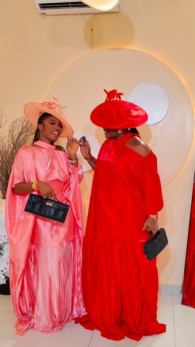 Two women in bright pink and red satin dresses wearing large, elegant statement hats.