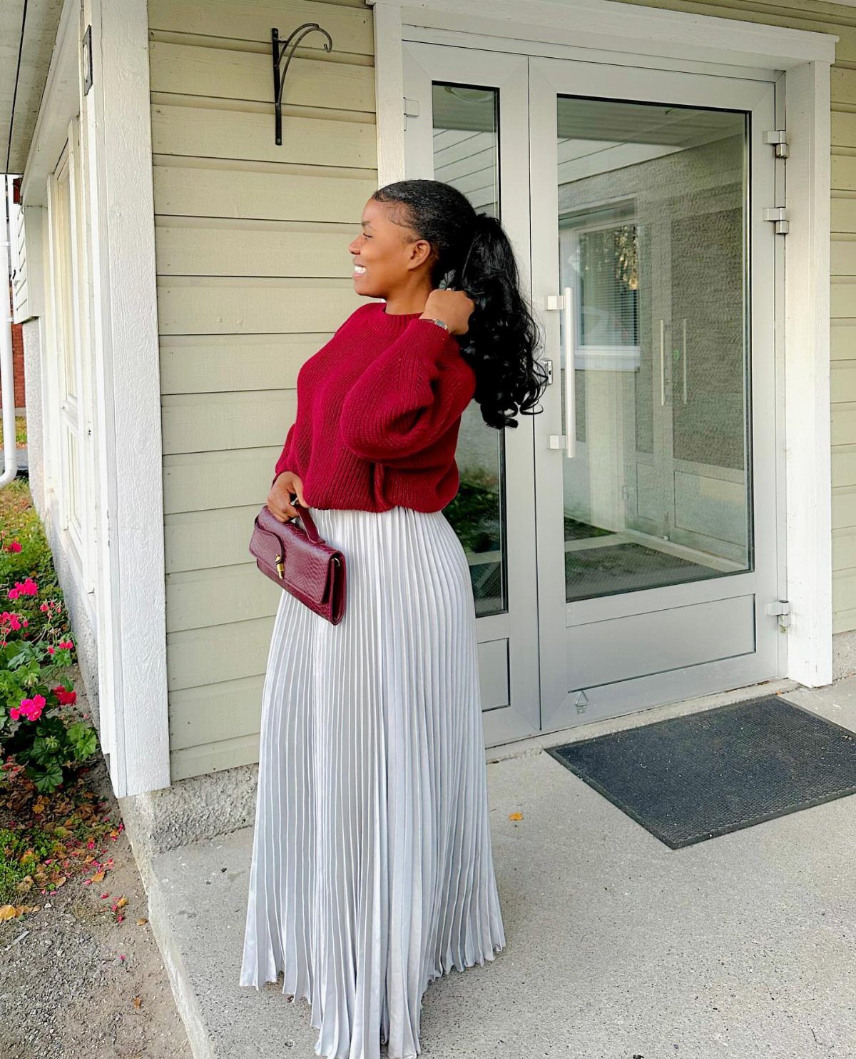 A woman wearing a red sweater and a long silver pleated skirt standing outside.