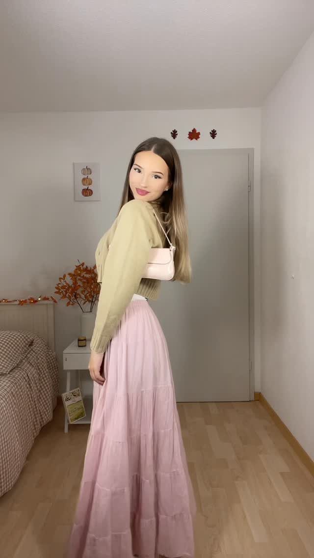 A young woman posing in a beige long-sleeved top and a long pink tiered skirt.