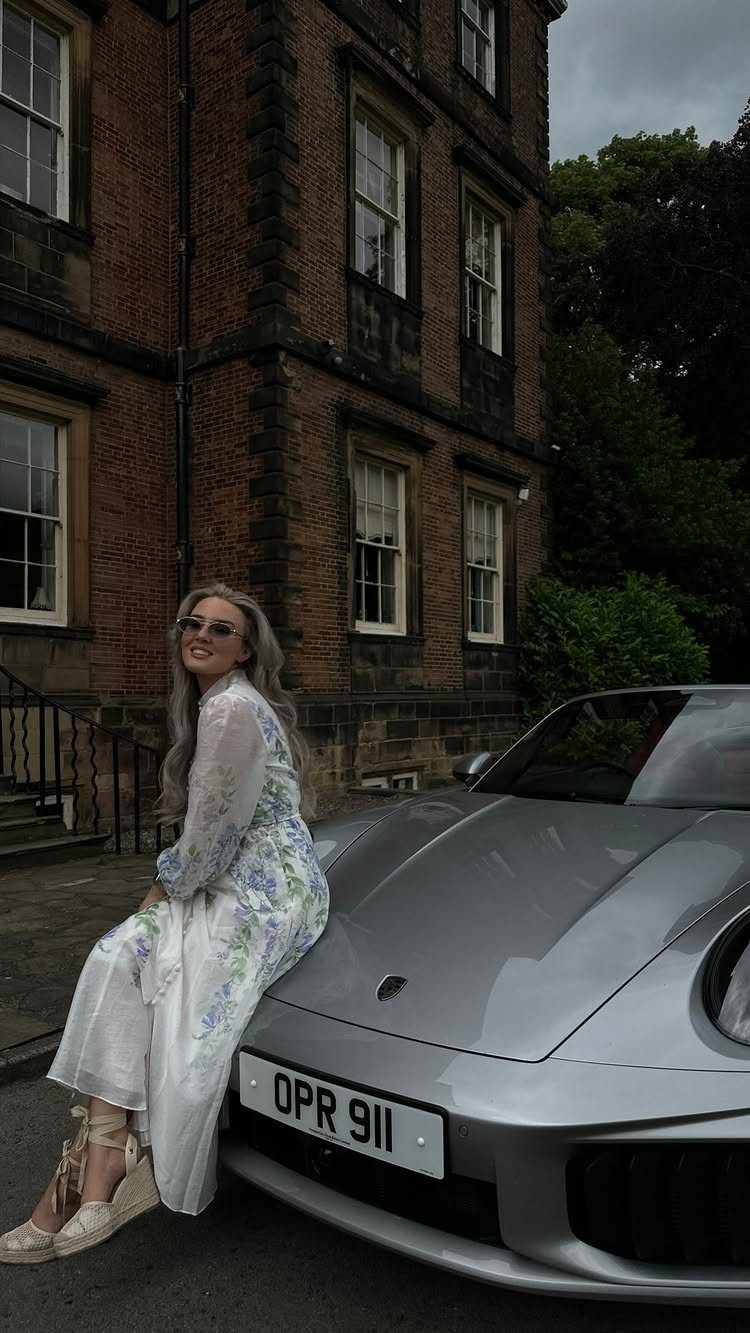 A woman in a white floral midi dress leaning against a silver sports car.