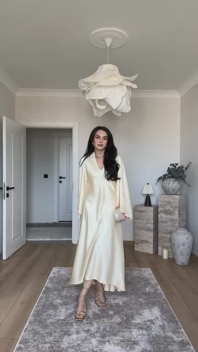 Woman in a cream long-sleeved maxi dress standing in a minimalist room with a floral light fixture.