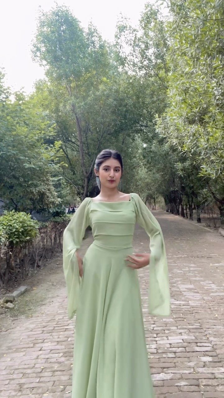 A woman in a long sage green dress with flared sleeves standing on a tree-lined path.