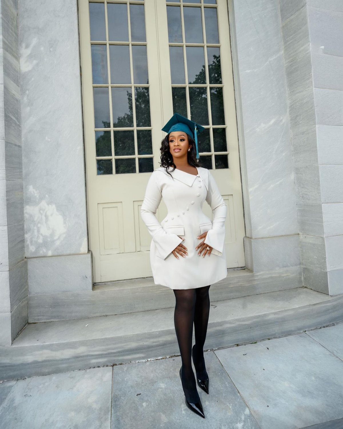 A woman in a white structured dress and teal graduation cap posing by a door.
