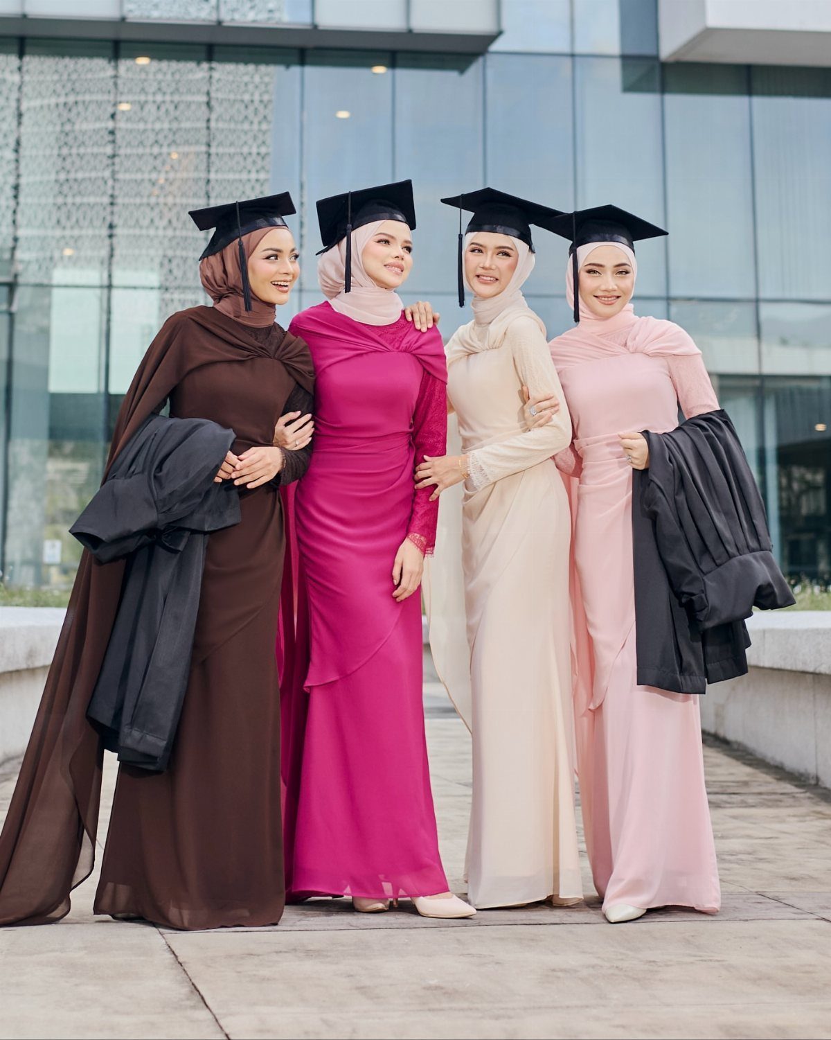 Four women in colorful modest graduation dresses and hijabs posing together outdoors.