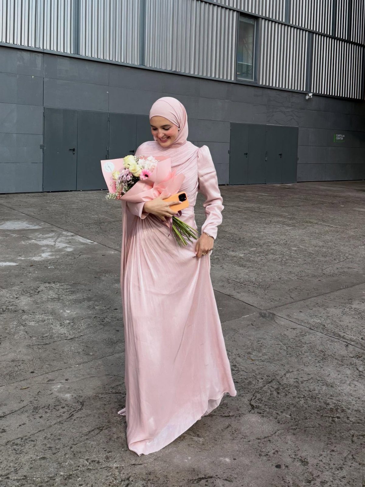 A woman in a light pink long-sleeved maxi dress and hijab holding flowers.