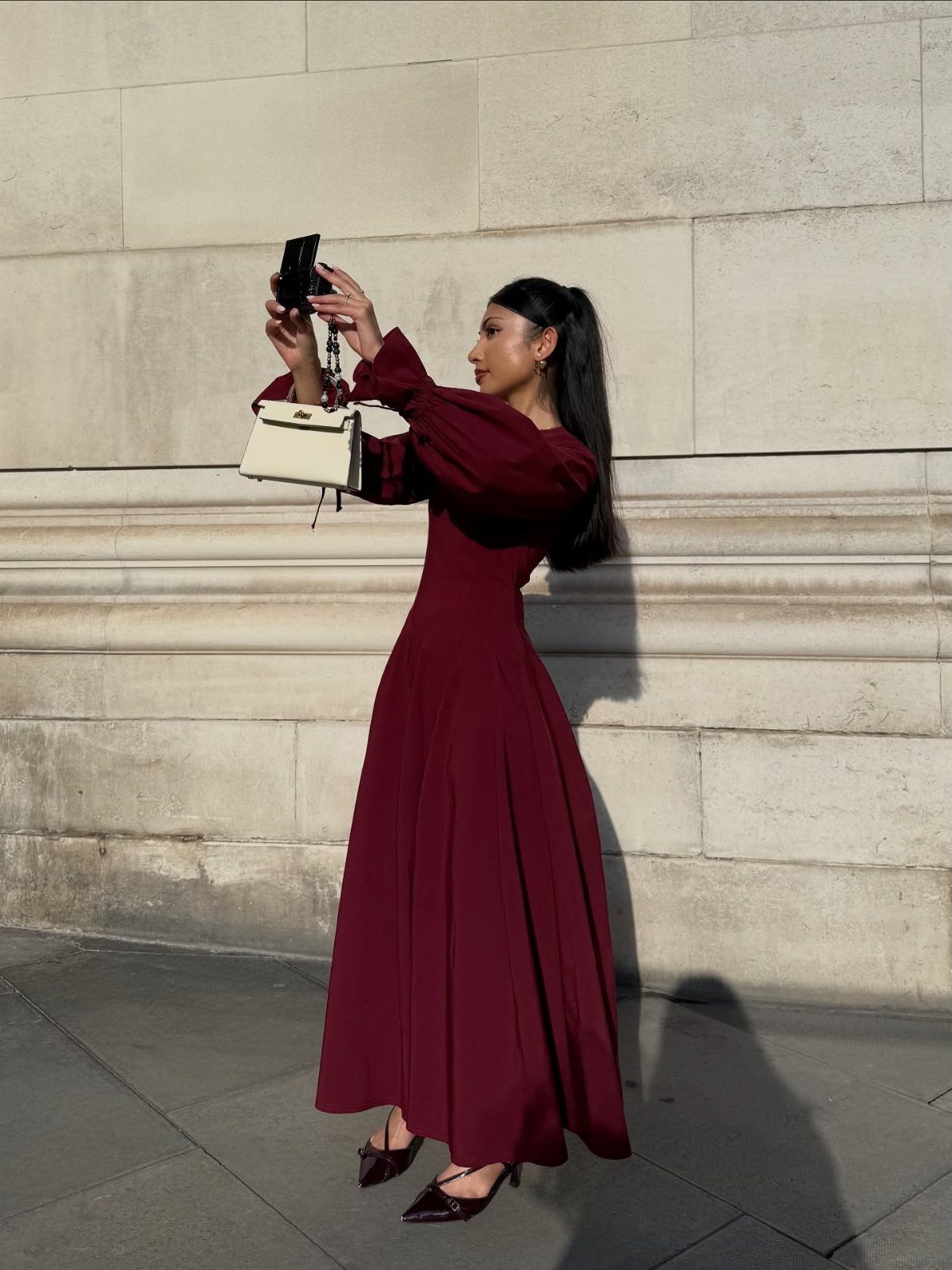 Woman in a long-sleeved maroon maxi dress taking a selfie against a stone wall.