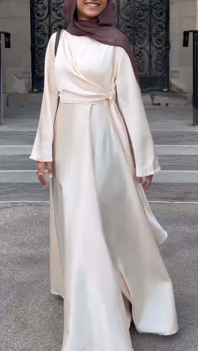 Woman in a long-sleeved cream satin maxi dress and brown hijab standing on stone steps.