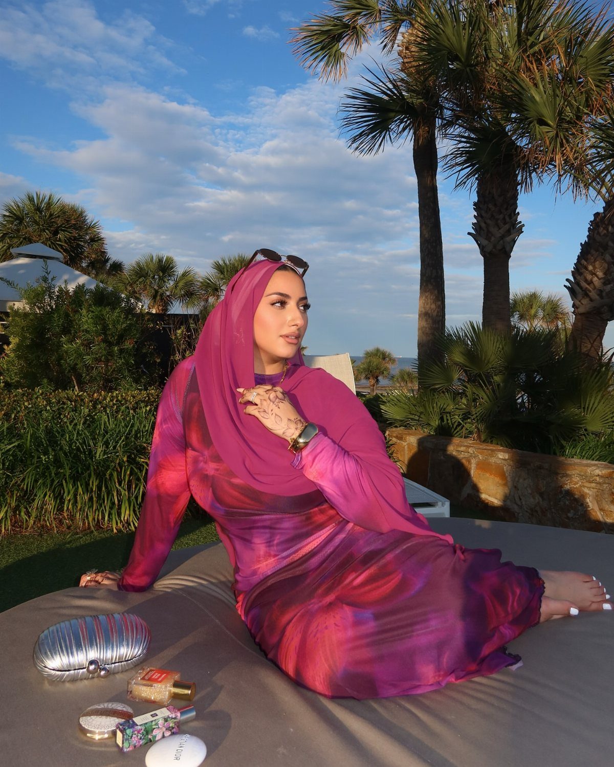 Woman in a flowing pink and purple chiffon dress and hijab sitting outdoors near palm trees.
