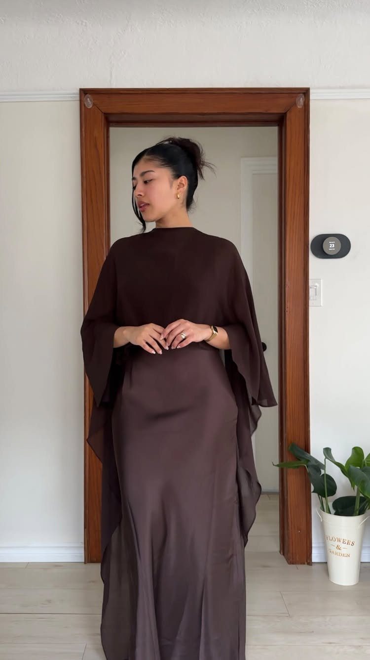 A woman wearing a long, flowing dark brown maxi dress with wide draped sleeves standing in a doorway.