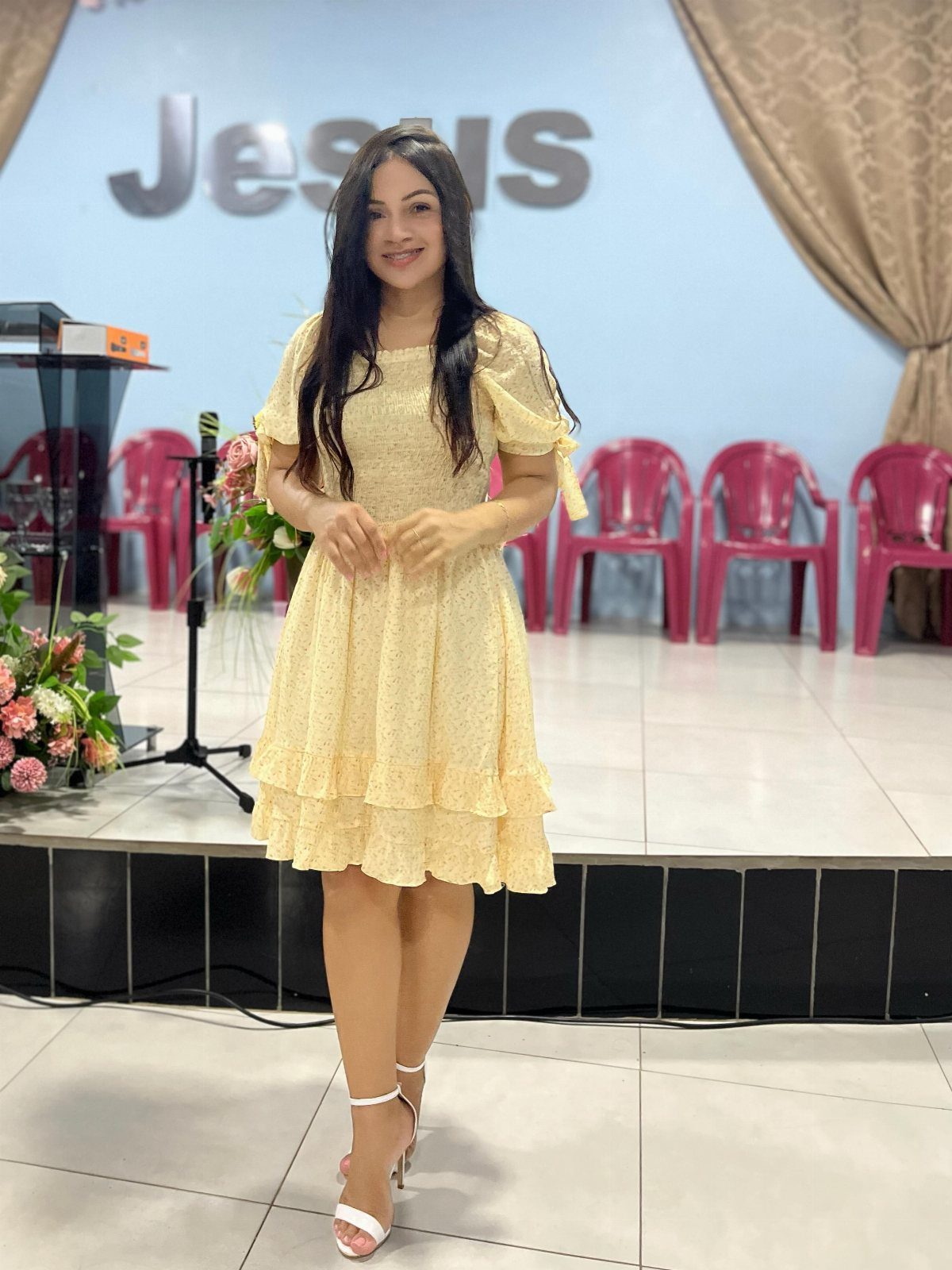 Smiling woman in a yellow lace dress standing on a stage with Jesus signage behind her.