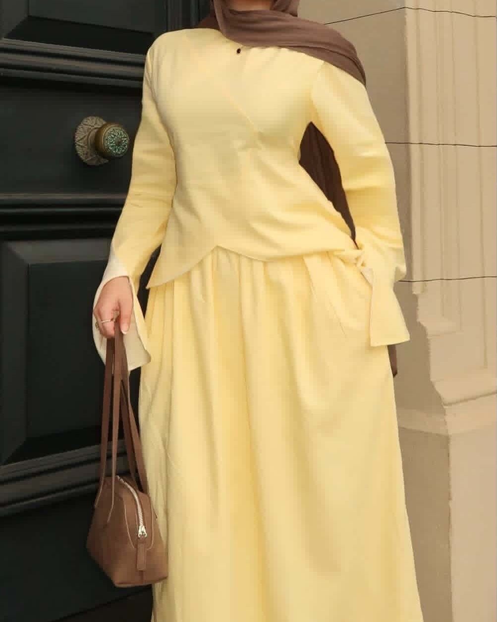 A person wearing a long-sleeved pale yellow maxi dress and a brown hijab holding a handbag.