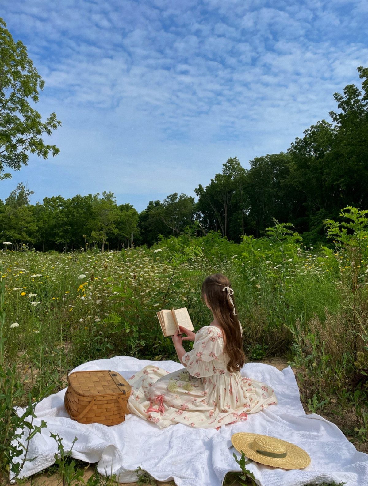 A girl in a floral dress reads a book while sitting on a picnic blanket in a meadow.