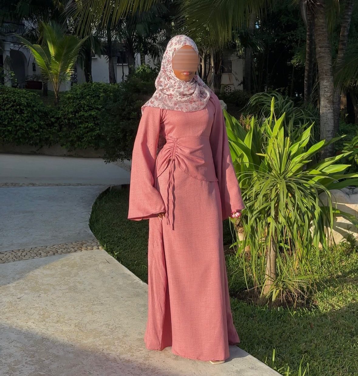 Woman in a long-sleeved dusty rose maxi dress and patterned hijab standing in a tropical garden.