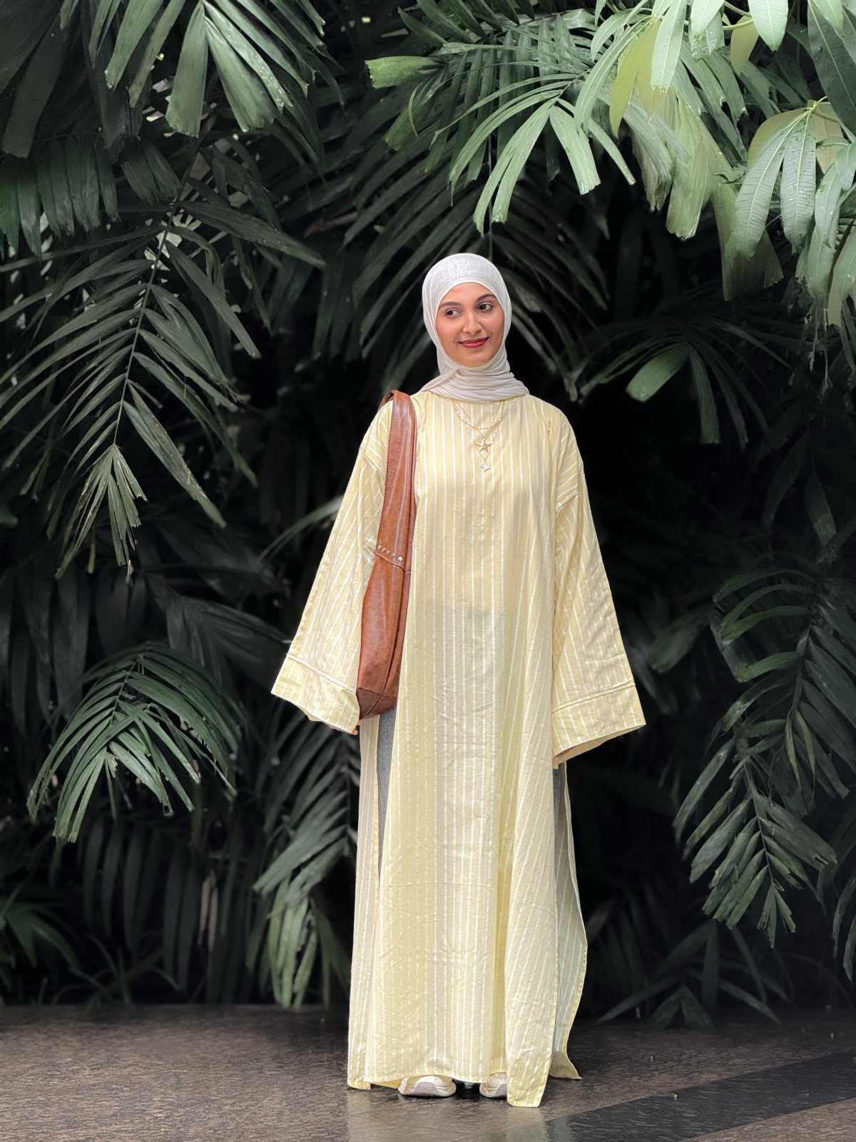 A woman in a long yellow striped dress and white hijab standing before tropical leaves.
