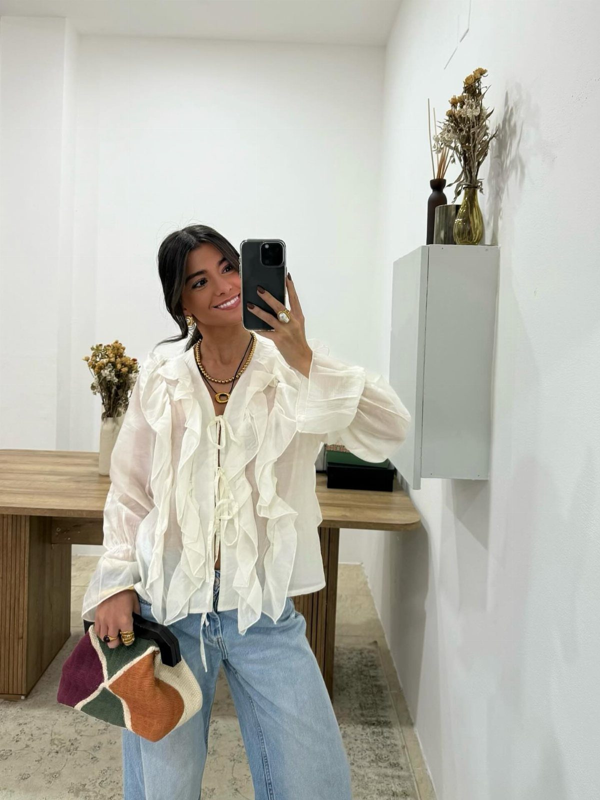 Woman in a white ruffled blouse and blue jeans taking a mirror selfie.