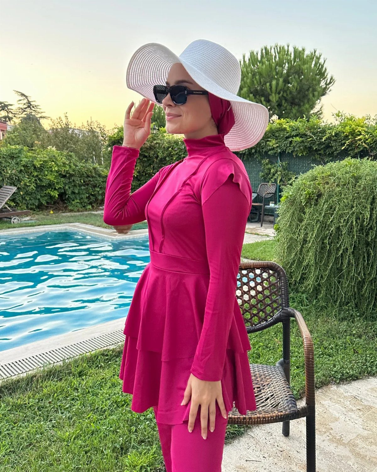 Woman in pink modest swimwear and white sun hat standing by a swimming pool.