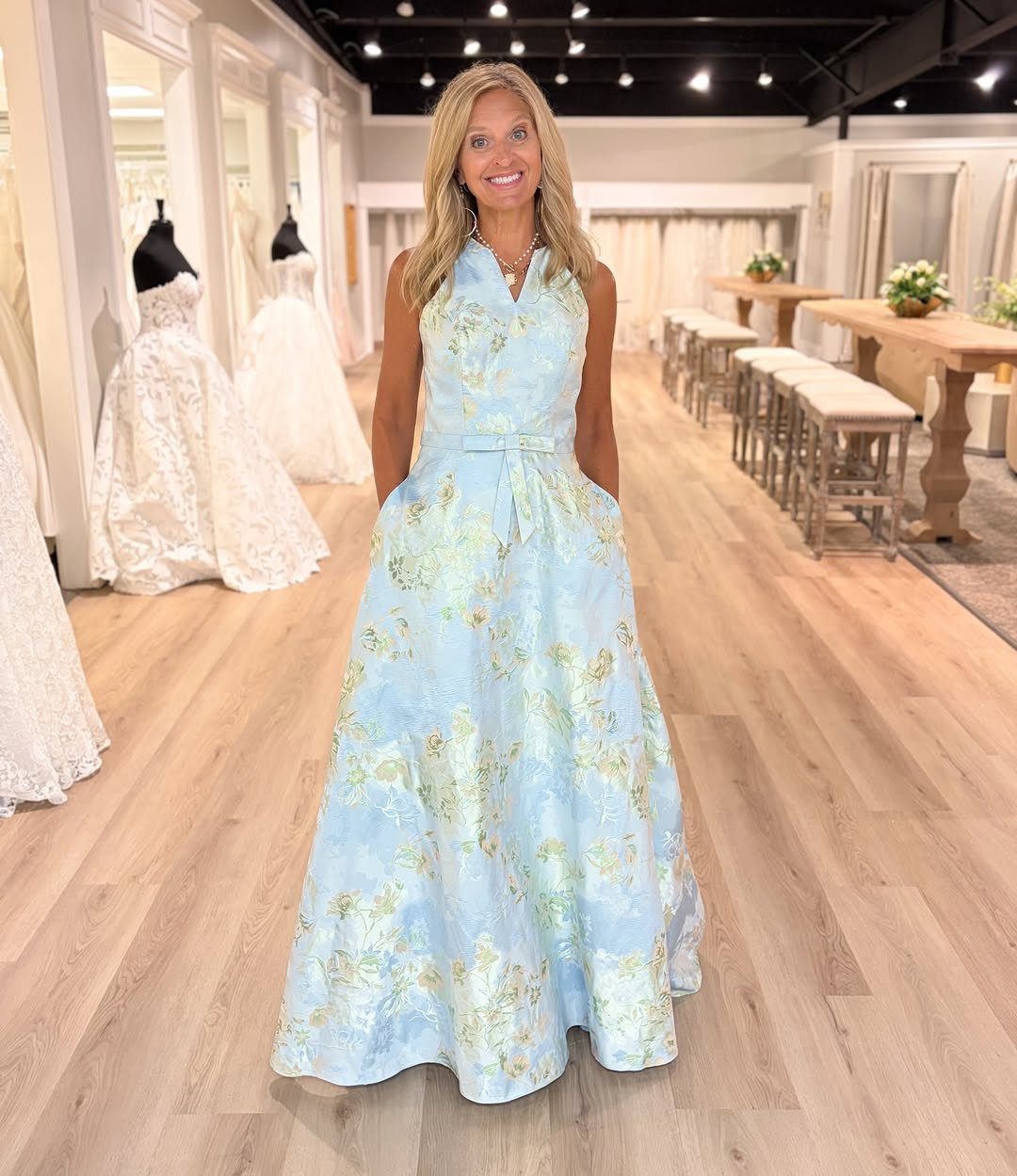 A woman smiling while wearing a light blue sleeveless dress with large floral patterns.