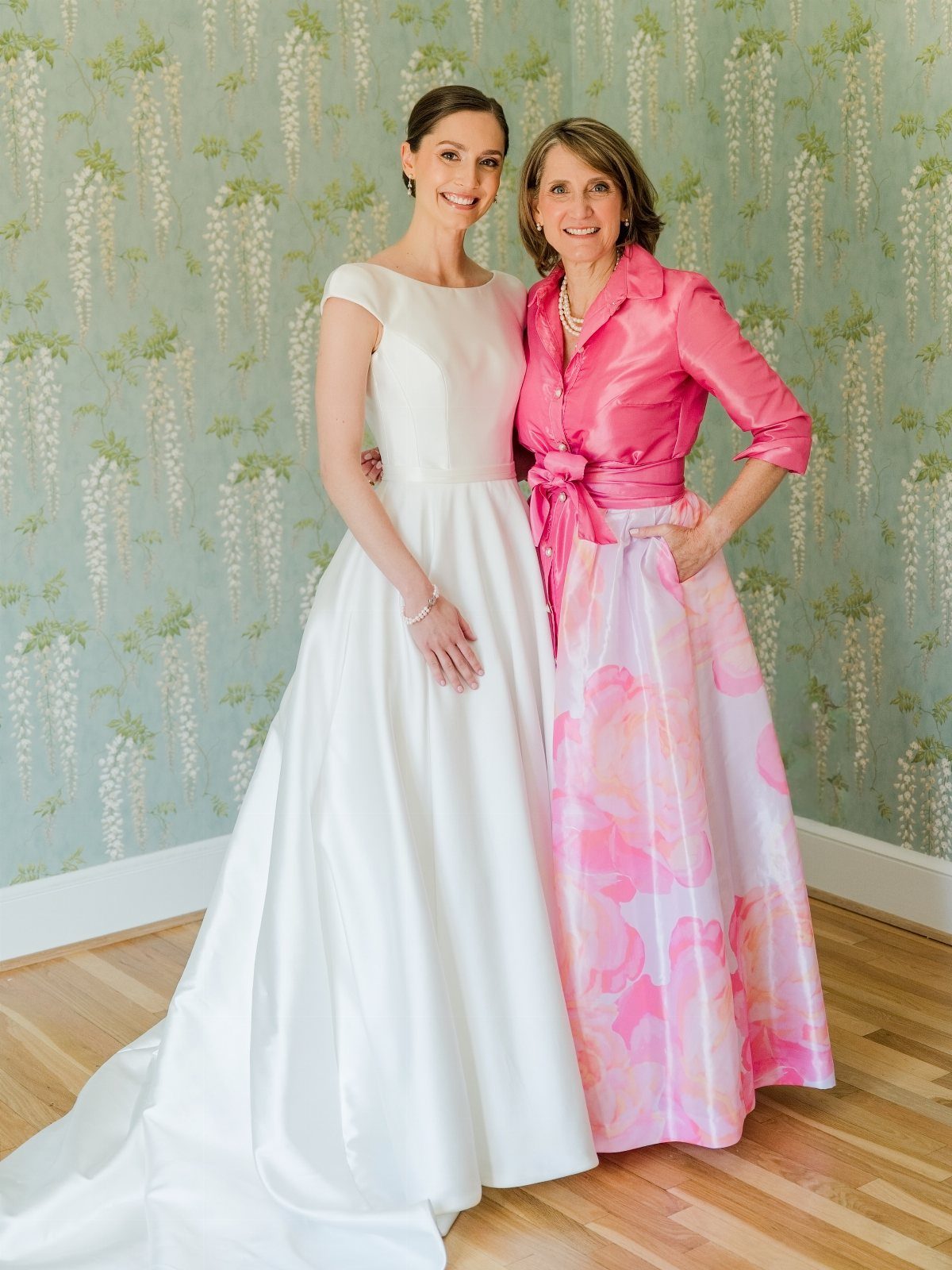 A bride in white and a woman in a pink floral skirt posing together.