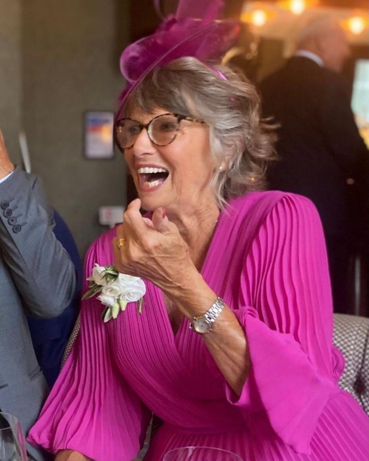 A laughing older woman wearing a bright pink pleated dress and a matching fascinator.