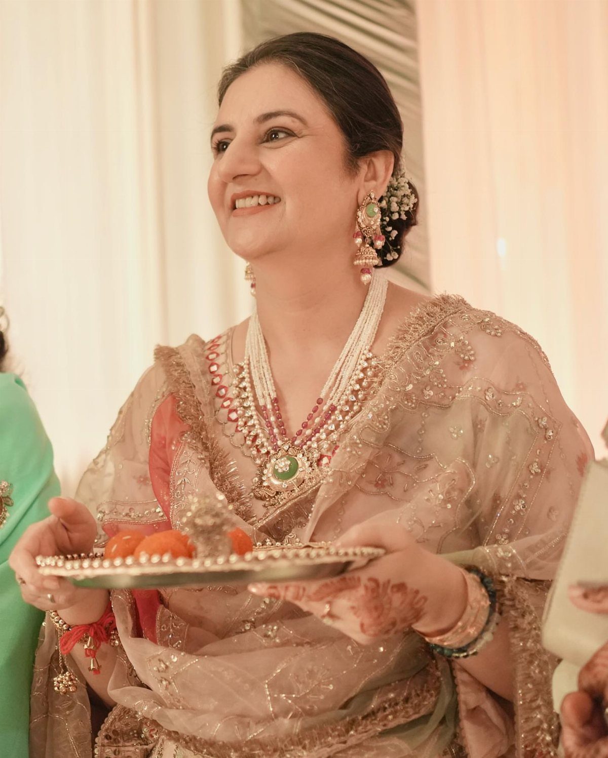 A smiling woman in an embroidered beige sari holding a silver tray with orange flowers.