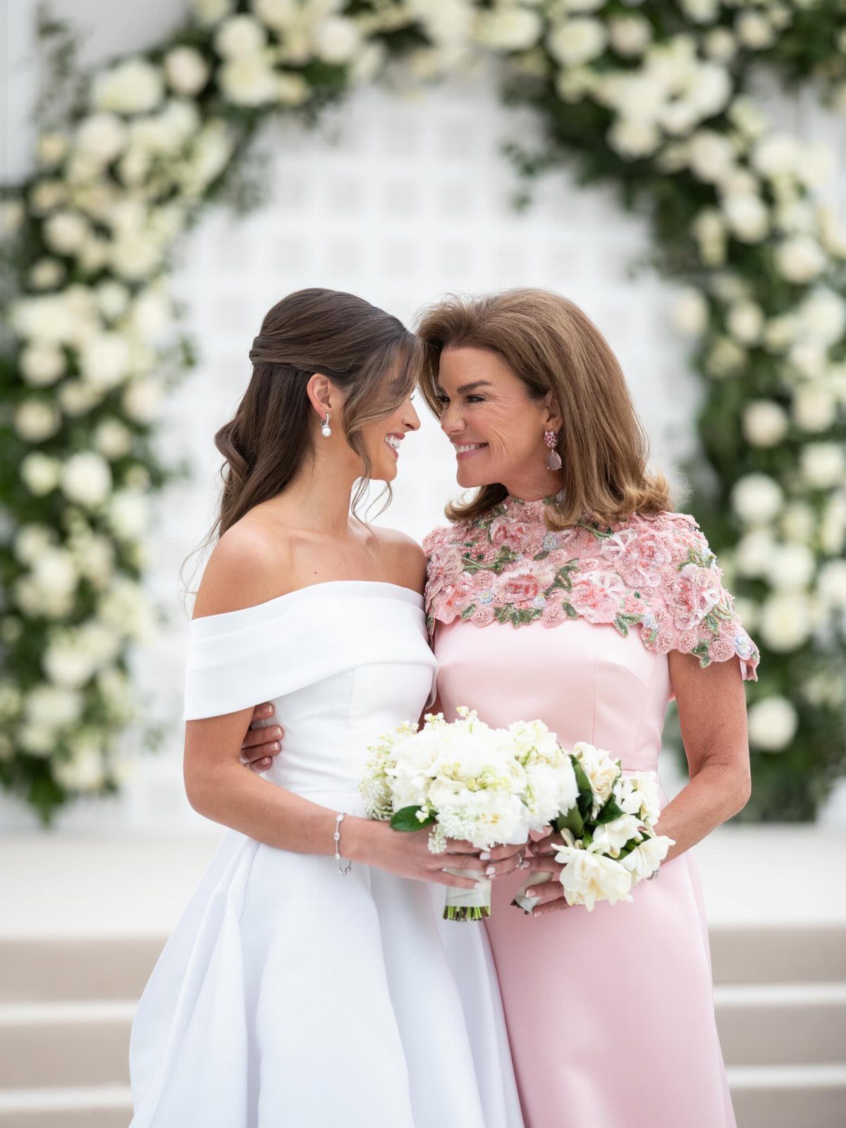 Two women smiling at each other in front of a large white floral wedding arch.