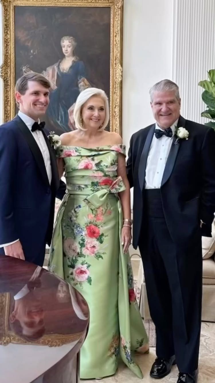 Three people in formal attire posing together in a room with a large portrait.