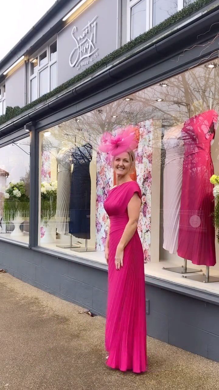 A woman in a bright pink formal gown and matching fascinator stands outside a boutique.