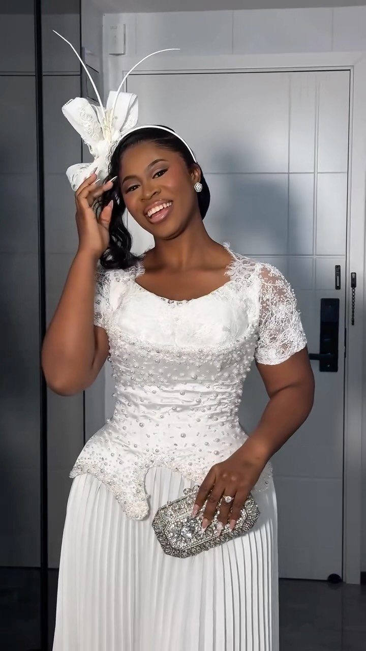 Smiling woman in a white lace wedding dress with a long pleated skirt and decorative headband.