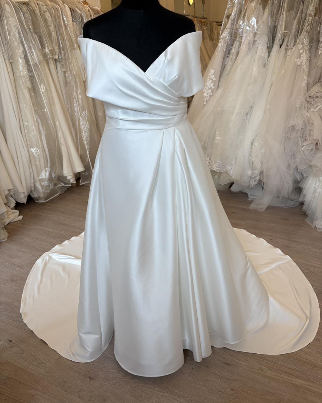 White satin off-the-shoulder wedding dress with a pleated bodice displayed on a mannequin.