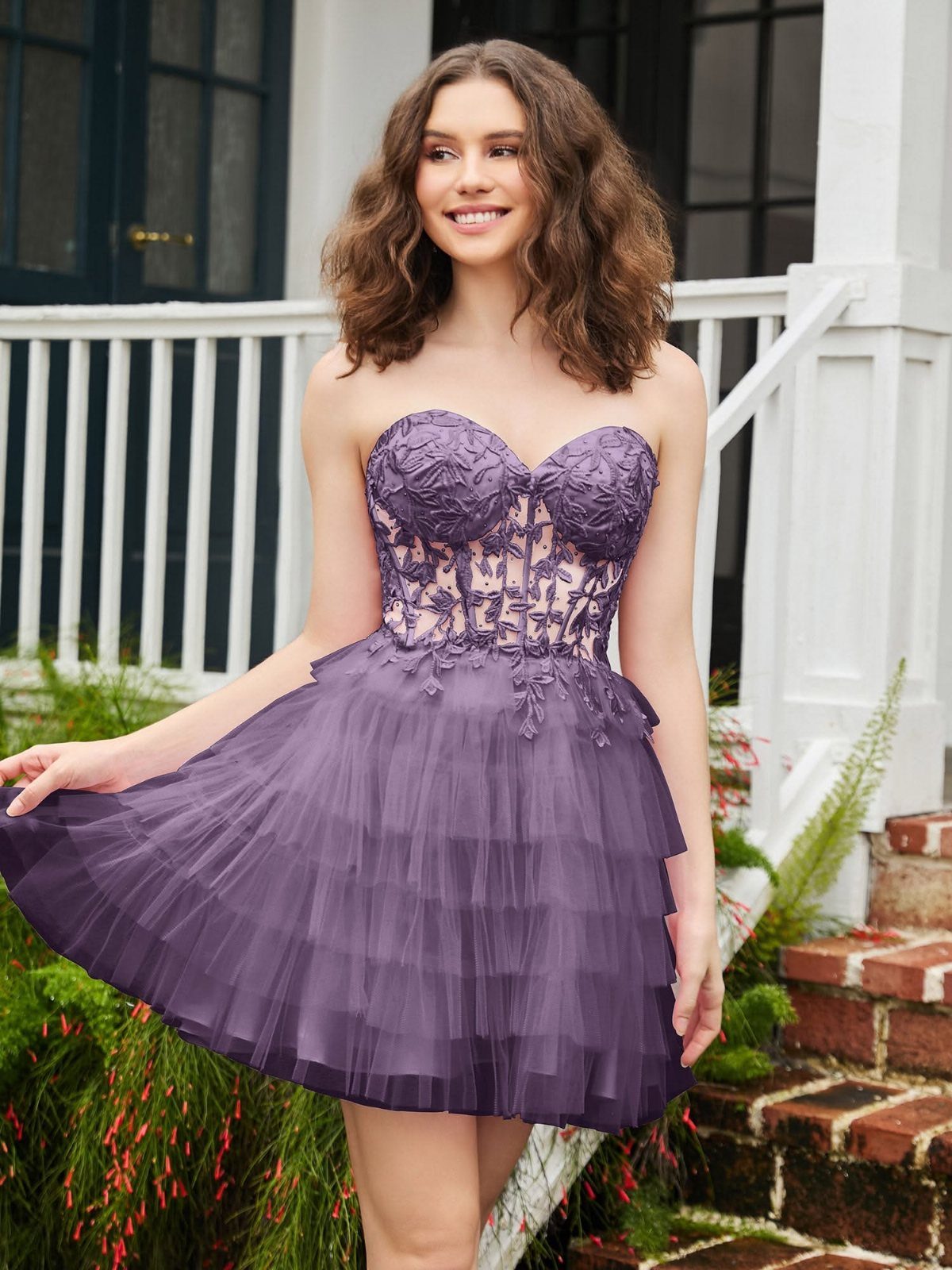 A smiling young woman wearing a plum-colored corset mini dress with a ruffled tulle skirt.