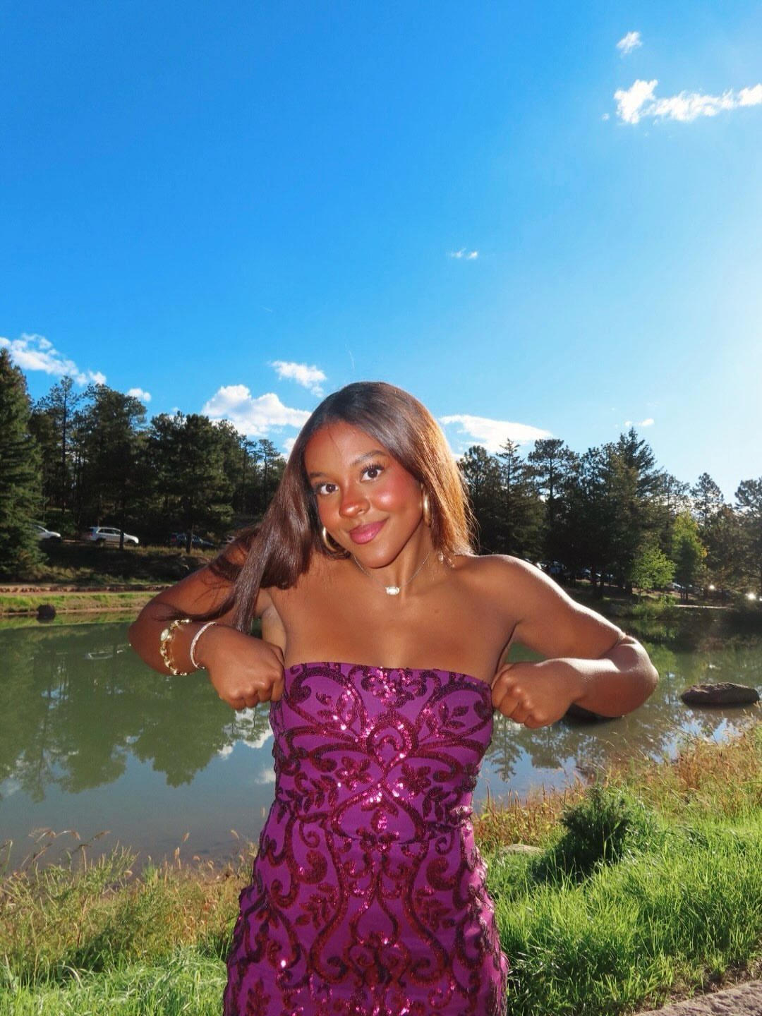 A young woman posing in a sequined plum strapless mini dress by a lake.
