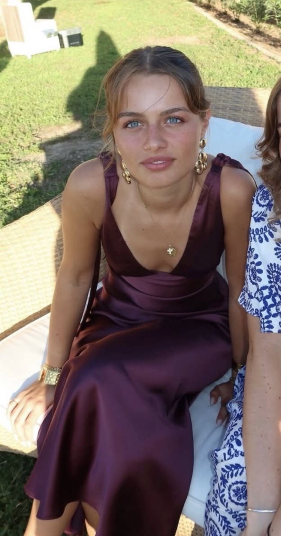Young woman wearing a plum satin slip dress while sitting on a white lounge chair outdoors.