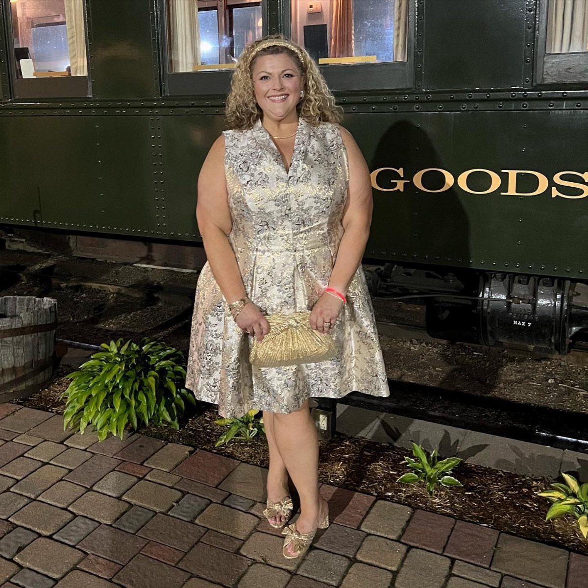 A woman in a gold brocade sleeveless dress posing in front of a green train car.