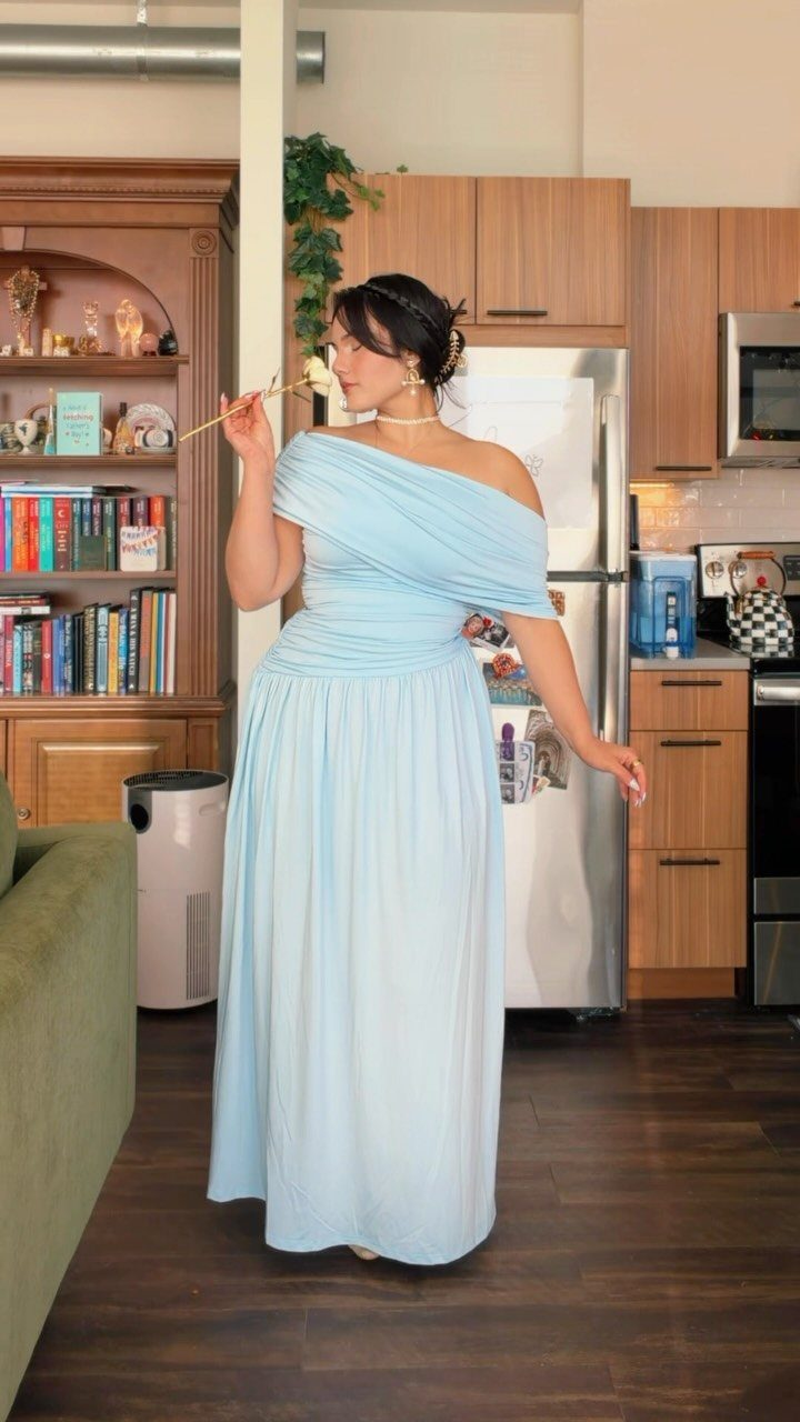 Woman in a light blue one-shoulder flowing maxi dress standing in a kitchen.