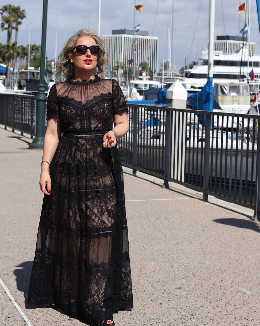 A woman in a black lace maxi dress walking along a sunny waterfront pier.