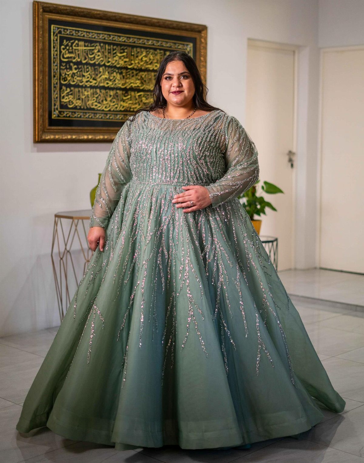 A plus size woman wearing a sage green sequined formal gown posing indoors.