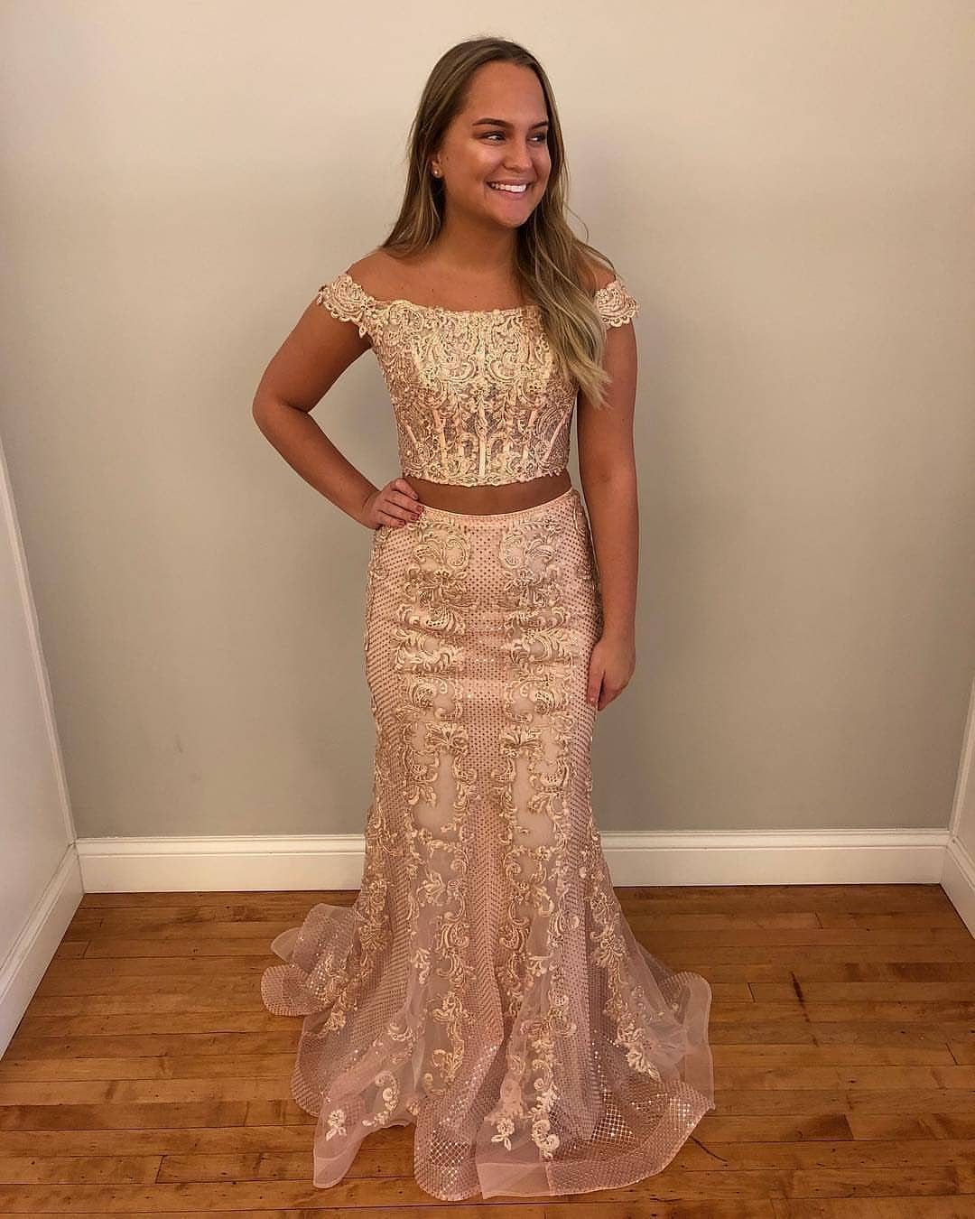 A smiling woman wearing a sparkling two-piece gold sequined prom dress against a plain wall.